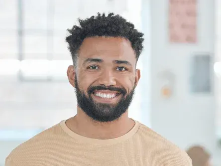 Smiling man with curly hair and beard wearing a tan sweater indoors.
